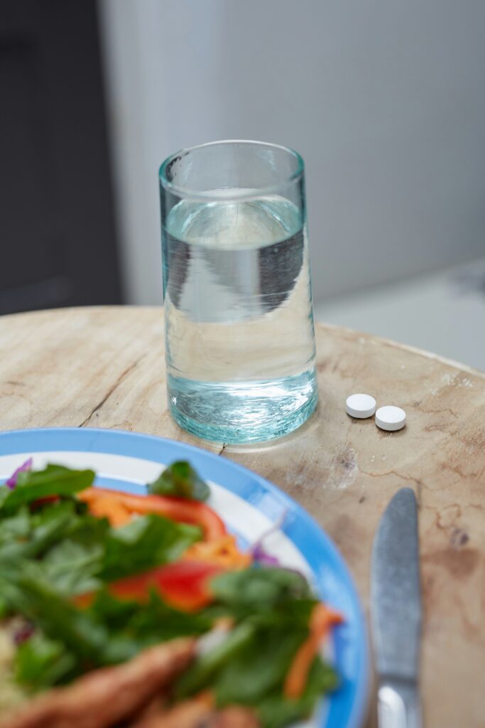 A glass of water and two white pills next to a healthy meal, representing medication during ramadan fasting
